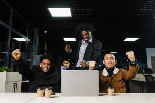 Happy and joyful multiethnic men raise their hands and fists and rejoice in winning in online casino on laptop. Team of black Africans and Asian programmer successful completed project