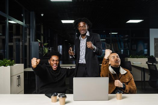 Emotional and joyful black Africans and Asians teamwork together and rejoice at end of online project. Multiethnic colleagues working on laptop, raising their hands, fists and shouting loudly