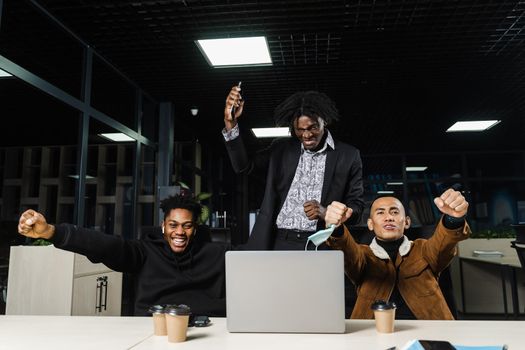 Happy and joyful multiethnic men raise their hands and fists and rejoice in winning in online casino on laptop. Team of black Africans and Asian programmer successful completed project