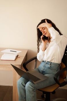 Depressed girl overtime working online with laptop on lap and talking by phone with colleagues. Exhausted overstressed attractive young woman feeling sick and having headache