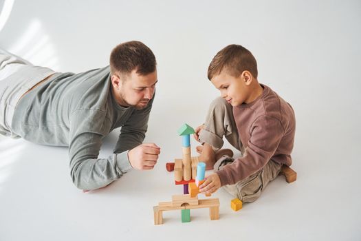 Caring dad helps his son to play on the floor on white background. Father and child build tower of colorful wooden bricks and have fun together