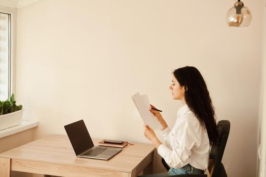 Attractive girl student studying online using laptop at home. Online education and distance learning. Girl holding papers in hands