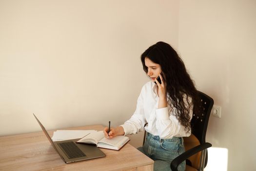 Busy woman talking by phone, write notes on notebook and working on laptop. Tired girl boss working online with colleagues at home