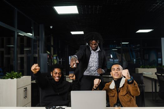 Emotional and joyful black Africans and Asians teamwork together and rejoice at end of online project. Multiethnic colleagues working on laptop, raising their hands, fists and shouting loudly