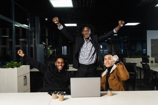 Happy and joyful multiethnic men raise their hands and fists and rejoice in winning in online casino on laptop. Teambuilding of black Africans and Asian programmers