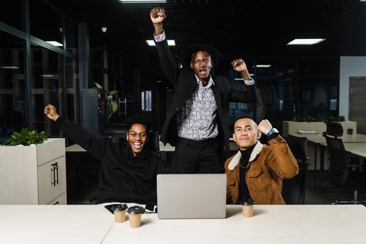 Happy and joyful multiethnic men raise their hands and fists and rejoice in winning in online casino on laptop. Teambuilding of black Africans and Asian programmers