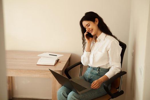 Cheerful woman manager with laptop on lap talking by phone, doing paperwork. Girl boss smiling and work in the office