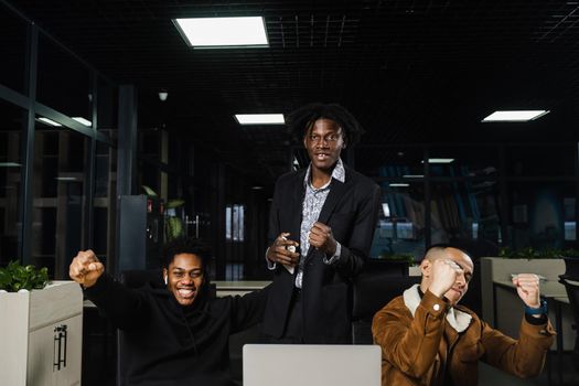 Emotional and joyful black Africans and Asians teamwork together and rejoice at end of online project. Multiethnic colleagues working on laptop, raising their hands, fists and shouting loudly