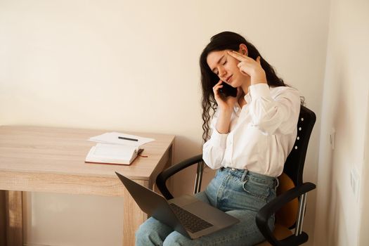 Depressed girl overtime working online with laptop on lap and talking by phone with colleagues. Exhausted overstressed attractive young woman feeling sick and having headache