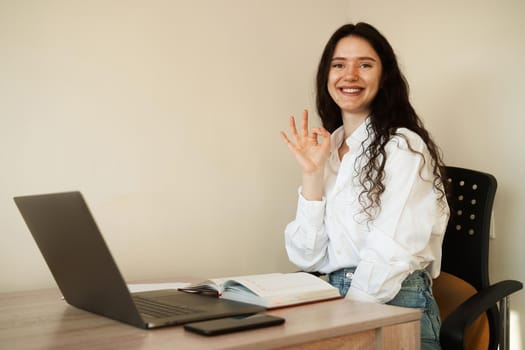 Schoolgirl with laptop done homework online and gesturing okay sign at table at home. Girl student showing ok sign.