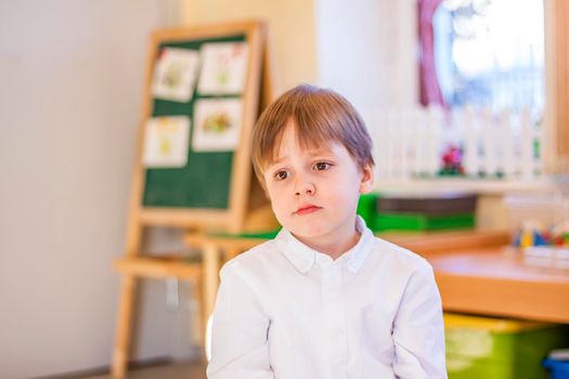 Elegantly dressed in a white shirt, a little boy is sitting in the classroom for lessons. portrait of a boy, blonde hair