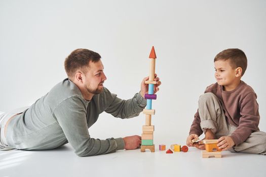 Caring dad helps his son to play on the floor on white background. Father and child build tower of colorful wooden bricks and have fun together