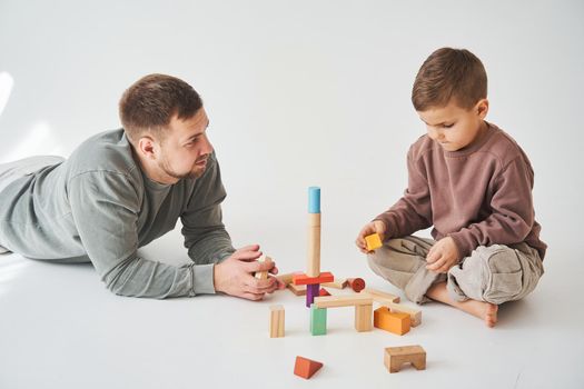 Caring dad plays with cheerful son with toy wooden cubes on white background. Fatherhood and child care