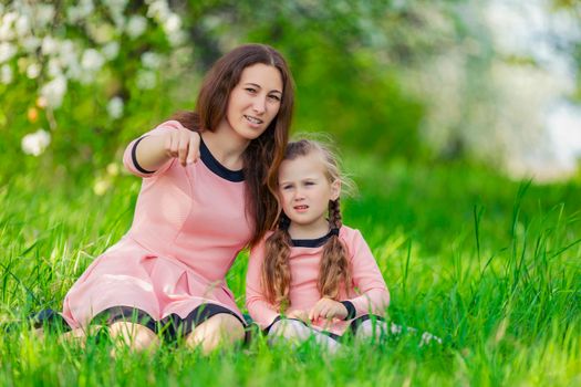 mother and daughter sit in the green grass against the backdrop of blooming apple trees