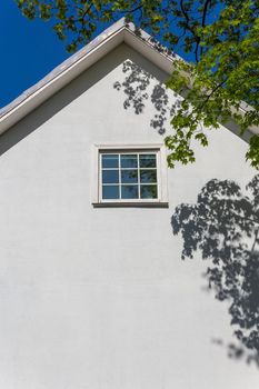 The white empty wall of a two-story old house with a gable roof on a sunny summer day. There are tree shadows on the wall. It can be used as a background for posting information.