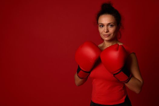 Martial art concept. Determined sportswoman, female athlete, boxer poses with boxing gloves against red background with copy space for advertising for boxing day event