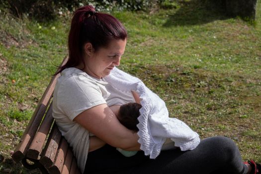 Woman is breastfeeding her child in a park, sitting on bench, hiding behind a stroller