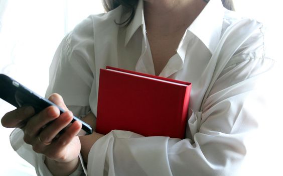 A beautiful girl in a white shirt holds a smartphone and a red book in her hands..