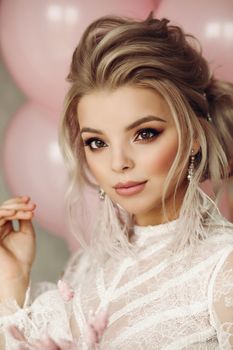 Fashion portrait of stunning blonde girl with hairstyle and professional make up in white transparent blouse holding bunch of pink colored flowers. Bunch of pink air balloons in background.