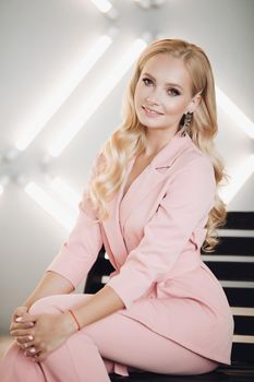 Attractive smiling blonde wearing pink smart suit sitting on stairs and posing. Young woman with long curls and makeup looking at camera on light background. Concept of outfit and beauty.