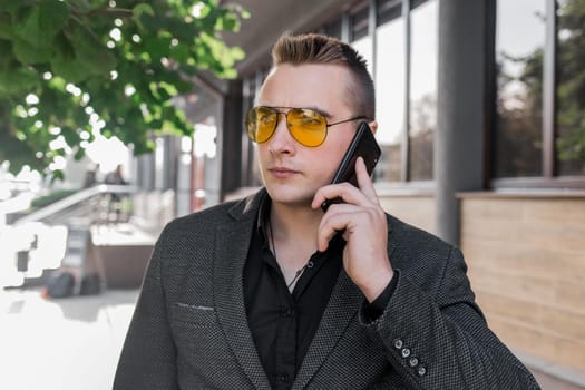 Stylish young businessman of attractive European appearance in sunglasses, jacket and shirt, talking on a mobile phone on the outdoor street.