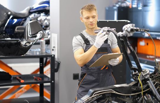 Portrait of maintenance service worker in uniform examine motorbike, man with clipboard paper. Pit stop, garage, high quality help, fixing station concept