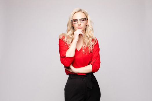 Beautiful young businesswoman in glasses thinking seriously and posing at camera. Blonde pretty lady leaning one hand one other and touching face. Confident girl in red blouse looking thoughtfully.