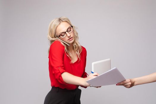 Side view of busy businesswoman talking by phone, using computer and signing papers in studio. Serious blonde looking at camera and working on isolated background. Concept of job and success.