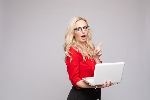 Front view of shocked woman wearing official clothes and glasses keeping computer. Amazed young businesswoman with open mouth looking at camera on isolated background. Concept of surprise.