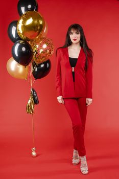 Young brunette model with blue eyes posing in fashionable clothes. Beautiful girl with long hair in elegant red costume looking at camera. Gorgeous lady standing near bunch of black and gold balloons.