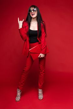 Side view of glamorous brunette posing in red smart suit and heels on red isolated background in studio. Young woman keeping glasses in hand and looking down. Concept of beauty and fashion.