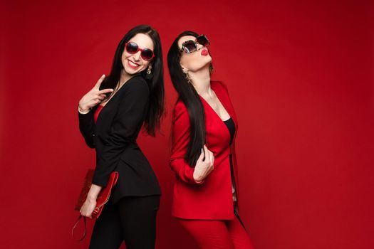 Two young fashion stylish female model in sunglasses smiling posing isolated at red studio background. Adorable happy woman friend relaxing having fun together medium long shot