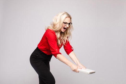 Side view of angry female manager crashing computer and shouting on white isolated background in studio. Furious woman in glasses irritating while working. Concept of problems and troubles.