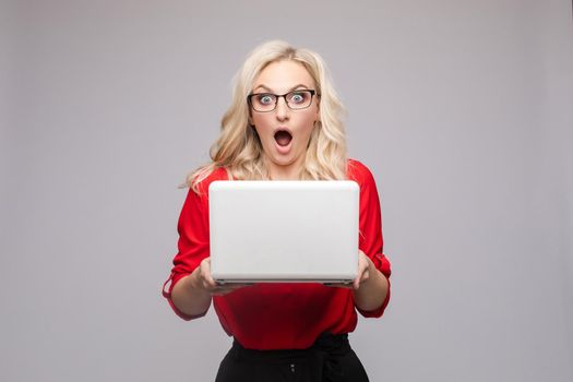Front view of shocked woman wearing official clothes and glasses keeping computer. Amazed young businesswoman with open mouth looking at camera on isolated background. Concept of surprise.