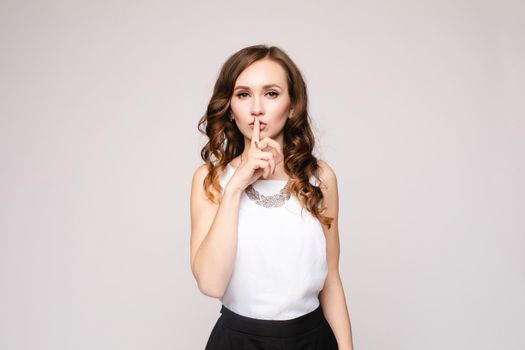 Beautiful pretty lady in white blouse decorated with silver accessory and black skirt posing at camera. Brunette woman holding finger near lips and asking being quite. Young girl not telling secrets.