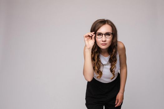 Front view of shocked long haired woman keeping glasses and looking with big eyes in camera. Young female in white shirt standing and posing on grey isolated background. Concept of surprise.