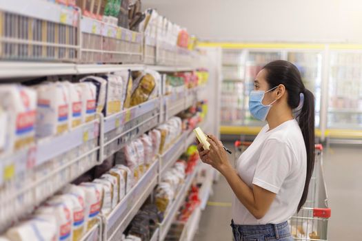 Asian woman wear face mask push shopping cart in supermarket. Girl looking grocery to buy something During coronavirus crisis or covid19 outbreak. Women checking her shopping items on list new normal concept