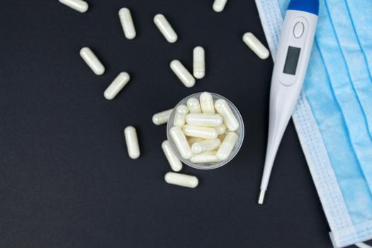 White electronic thermometer with packs of tablets and blue medical face mask on black background. Selective focus.