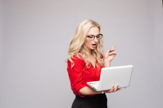 Front view of shocked woman wearing official clothes and glasses keeping computer. Amazed young businesswoman with open mouth looking at camera on isolated background. Concept of surprise.