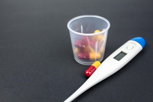 Medical capsules, tablets and clinical thermometer inside measuring cup on black background. Side view