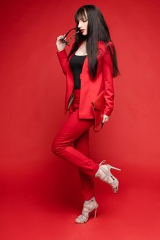 Side view of glamorous brunette posing in red smart suit and heels on red isolated background in studio. Young woman keeping glasses in hand and looking down. Concept of beauty and fashion.