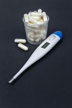 Medical capsules, tablets and clinical thermometer inside measuring cup on black background. Side view