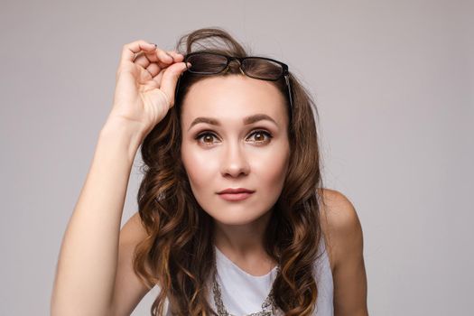 Front view of shocked long haired woman keeping glasses and looking with big eyes in camera. Young female in white shirt standing and posing on grey isolated background. Concept of surprise.