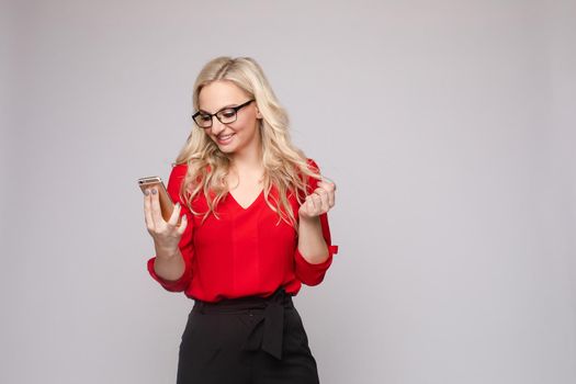 Front view of smiling woman wearing red blouse and skirt keeping phone and laughing on isolated background. Cheerful blonde messaging and communicating in studio. Concept of technology and business.
