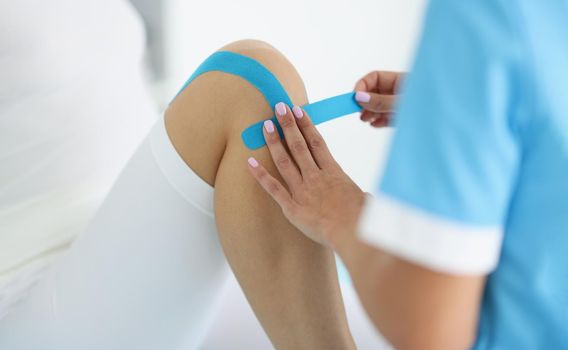 Close-up of physiotherapist put kinesiology tape on clients knee, remove pain, rehabilitation center, massage cabinet. Physical therapy, recovery concept