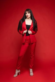 Side view of glamorous brunette posing in red smart suit and heels on red isolated background in studio. Young woman keeping glasses in hand and looking down. Concept of beauty and fashion.