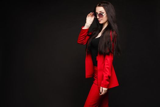 View from side of stylish brunette wearing glasses and red smart suit posing on black isolated background. Young lady looking at camera and smiling in studio. Concept of glamour and beauty.