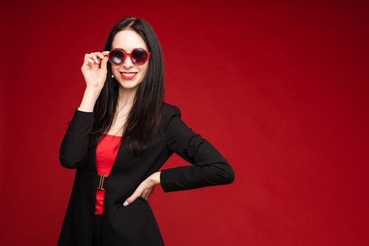 Portrait of young beautiful woman thoughtfully looking down. Brunette model in elegant costume posing at camera . Pretty lady in red sunglasses leaning one hand on other and holding it near face.