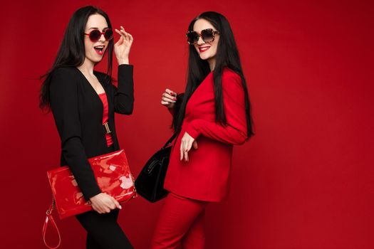 Side view of two brunettes wearing black and red smart suits and glasses standing back to back on isolated background. Stylish women posing and looking at camera in studio. Concept of glamour.