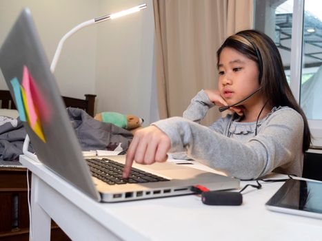  Asian girl in headphones sit at desk study online on laptop. Kid wear headset handwriting in notebook learning using internet lessons on quarantine. Girl student learning virtual internet online class from school due to covid pandemic.